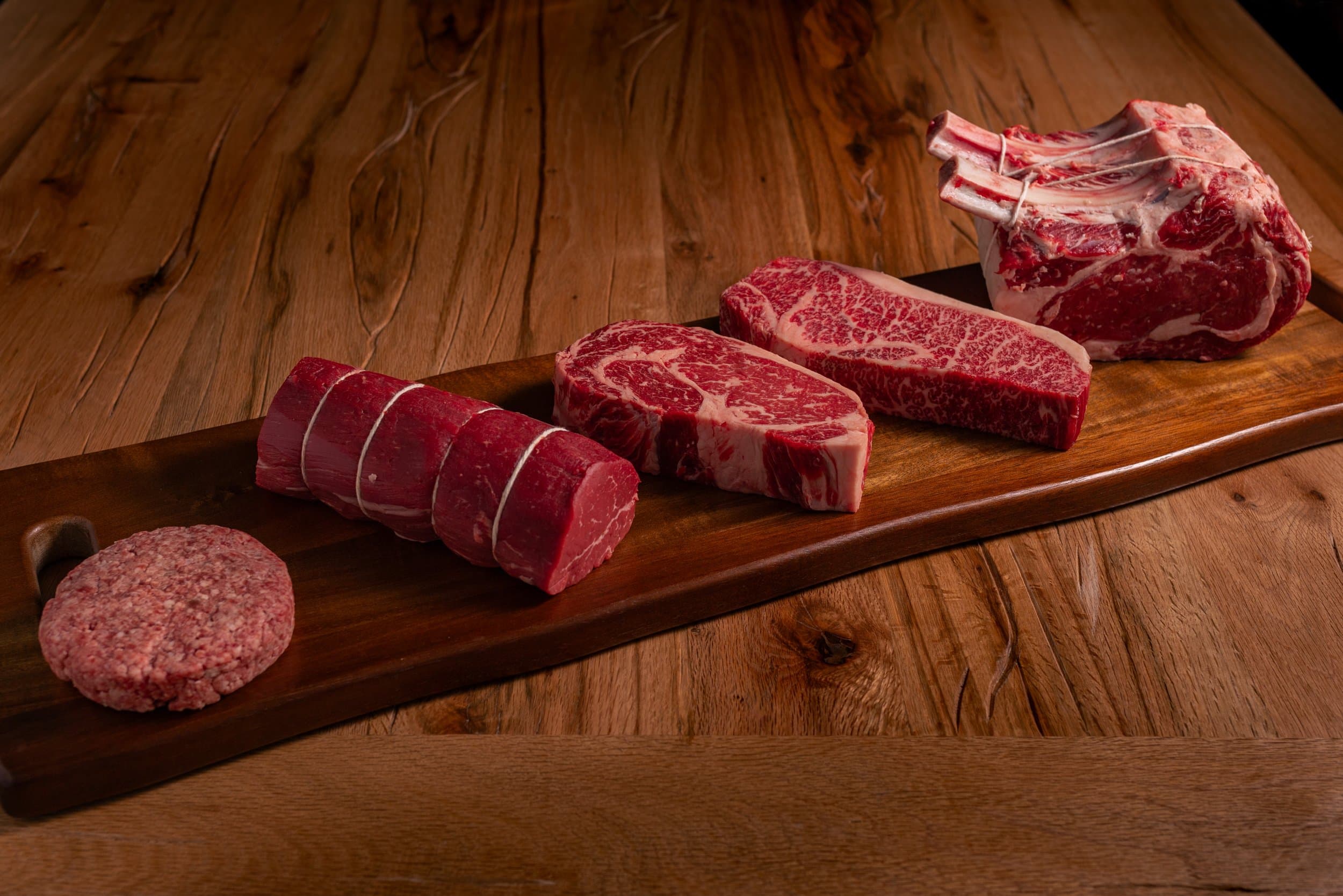 Ground beef patty, uncut filet, ribeye and Wagyu steaks and a bone-in prime rib roast on a wooden cutting board